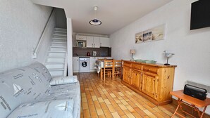 A compact living space with a kitchenette, a sofa with patterned cushions, a wooden dining table, and a small wooden cabinet. - Comfortable house, walk to beach and shops, pets allowed upon request (La Faute-sur-Mer)