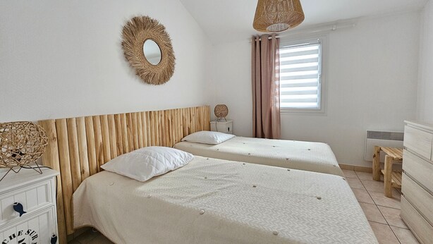 A bedroom with two beds, a wooden headboard, a mirror, a hanging light, and a window with curtains. - Comfortable house, walk to beach and shops, pets allowed upon request (La Faute-sur-Mer)