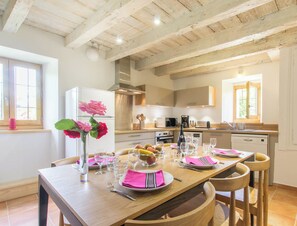 Una cocina moderna con vigas de madera, armarios blancos y una zona de comedor con una mesa de madera preparada para cuatro.