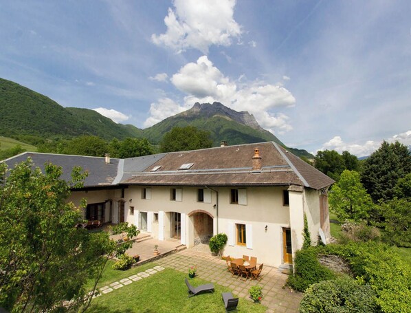 A traditional chalet with a stone patio, outdoor seating, and a mountain backdrop.