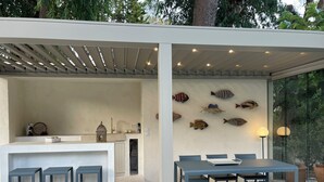 Ett modernt utomhusområde för måltider med ett vitt bord och blå stolar, en pergola med belysning och dekorativ väggkonst med fiskmotiv.