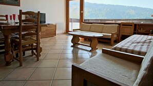 A cozy room with a wooden table, a sofa, and a TV. There's a large window with a mountain view. - Stunning 3-room apartment on top floor in Chamrousse (Chamrousse)
