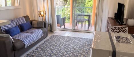 A living room with a sofa, a patterned rug, a television, and a balcony with outdoor furniture.