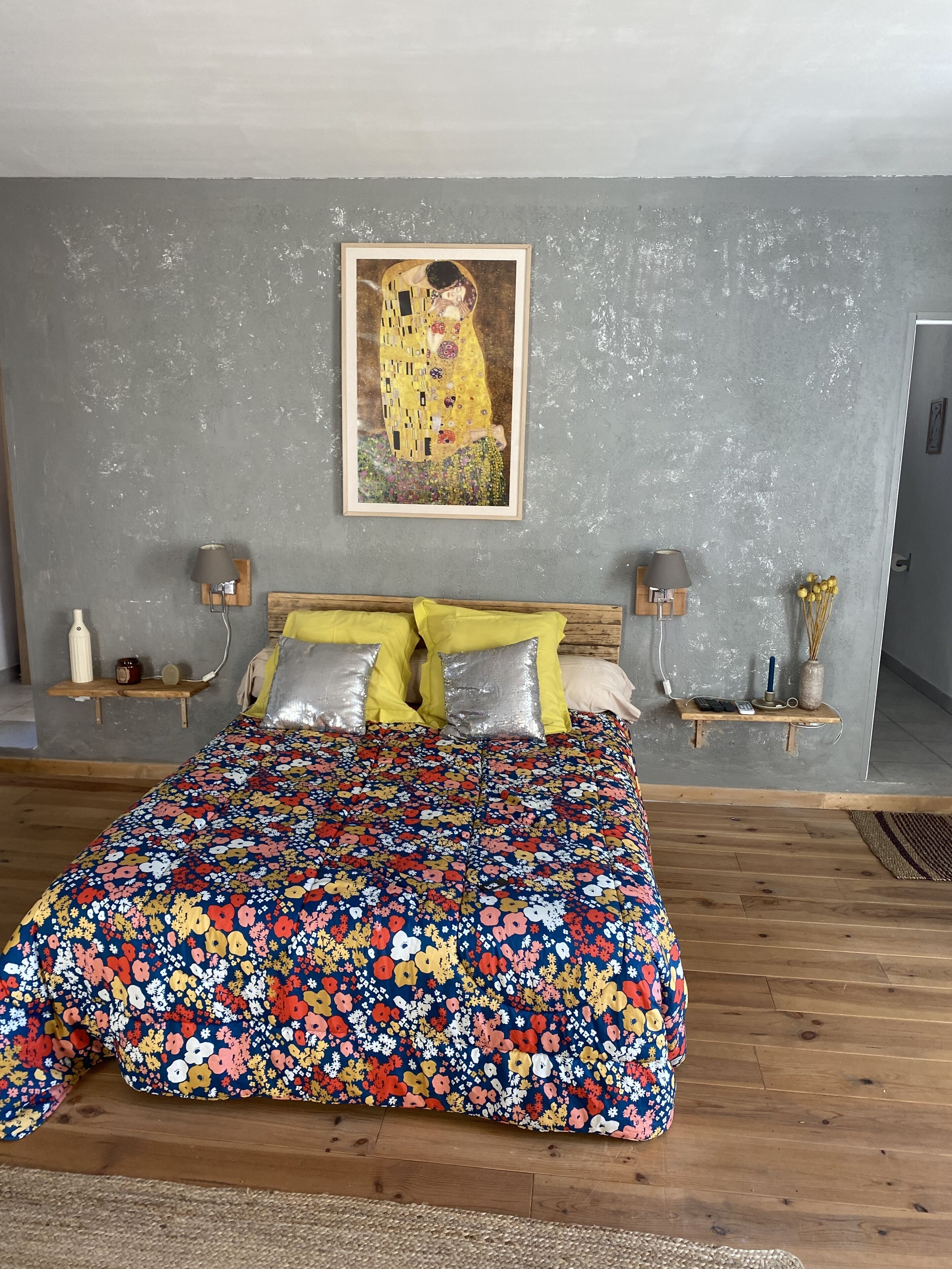 Une chambre à coucher avec une couvre-lit à motifs floraux, un grand tableau au mur et des tables de chevet ornées de lampes et de petits objets décoratifs.