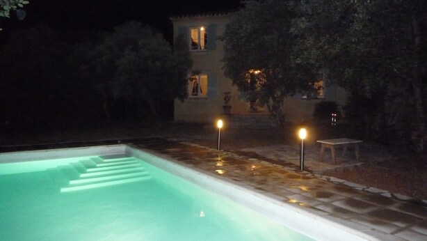 A swimming pool with a stone border, surrounded by a garden with trees and a building in the background. - Charming country house in the middle of the trees private swimming pool gorges de l'hérault  (Causse-De-La-Selle)