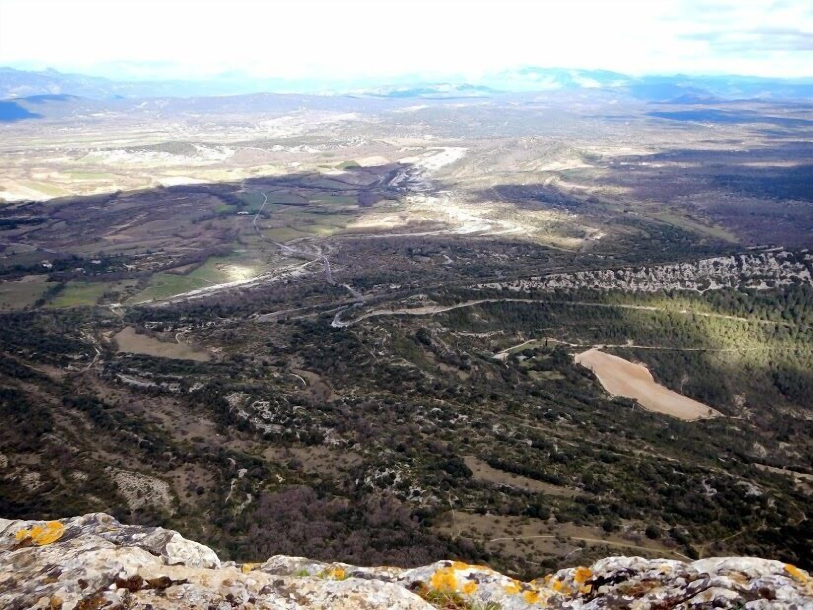 A panoramic view of a mountainous landscape with a winding road, patches of forest, and a small body of water.