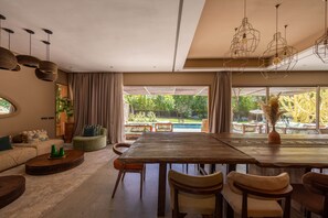 Een ruime woonkamer met een grote houten eettafel, comfortabele zithoek en grote ramen met uitzicht op een zwembad en tuin.