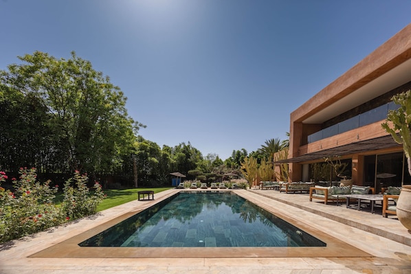 A rectangular swimming pool with a stone border, surrounded by a patio with outdoor furniture and a building with large windows.