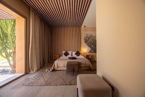 Een moderne slaapkamer met een bed, een bankje, een nachtkastje, een groot raam met gordijnen, en een cactus buiten.