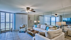 A spacious living room with a large sofa, a coffee table, and a chair. There are two ceiling fans, a large mirror, and a view of the ocean through the windows. - Steps from YOUR Balcony to Beach | Excellent Reviews | Beach Service Included (Miramar Beach)
