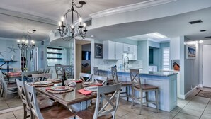 A modern kitchen with white cabinets, a central island, and a dining area with a wooden table and chairs. - Steps from YOUR Balcony to Beach | Excellent Reviews | Beach Service Included (Miramar Beach)