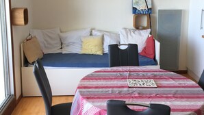 Ein gemütlicher Wohnraum mit Bett, Esstisch und Stühlen. Eine Lampe, ein Gemälde und ein Regal mit Gegenständen sind an der Wand angebracht.