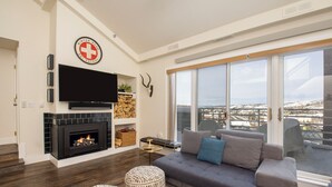 Una sala de estar moderna con chimenea, un sofá, otomanas y un amplio ventanal con vistas a la ciudad.