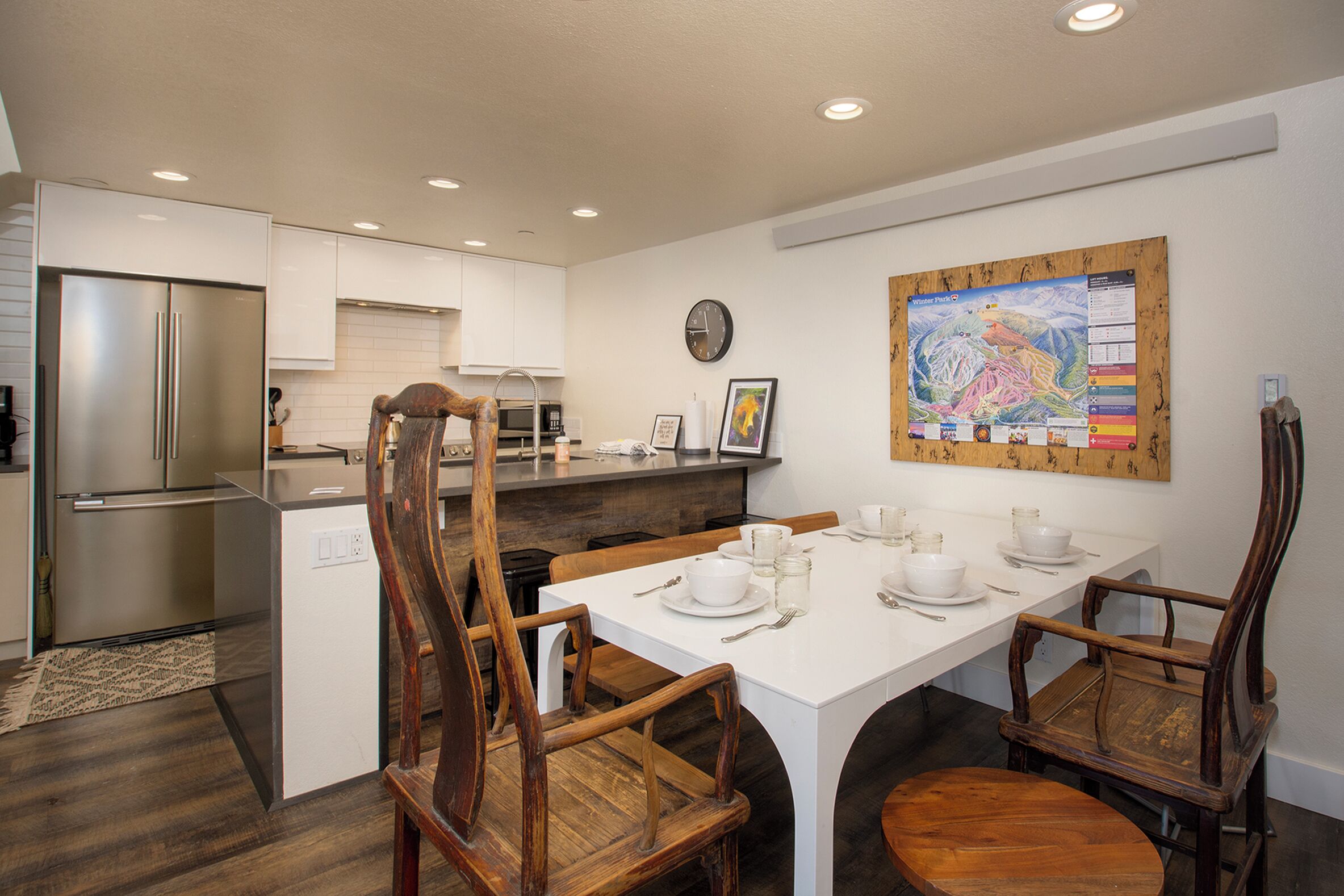 A modern kitchen with white cabinetry, a stainless steel refrigerator, wooden chairs, and a dining table set for four.