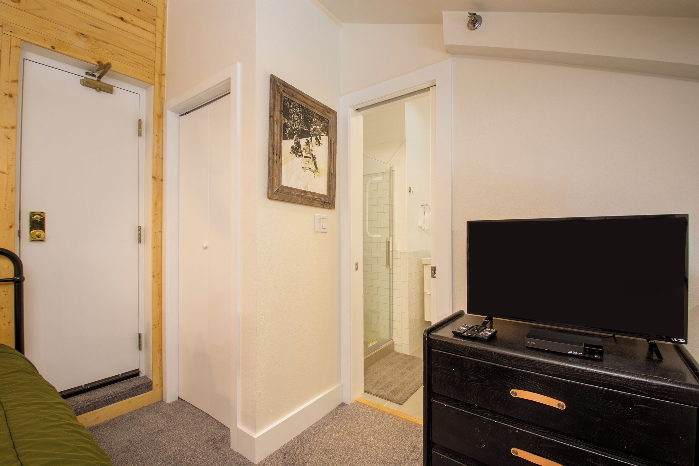 A bedroom with a bed, a dresser, a television, and a door leading to a bathroom.