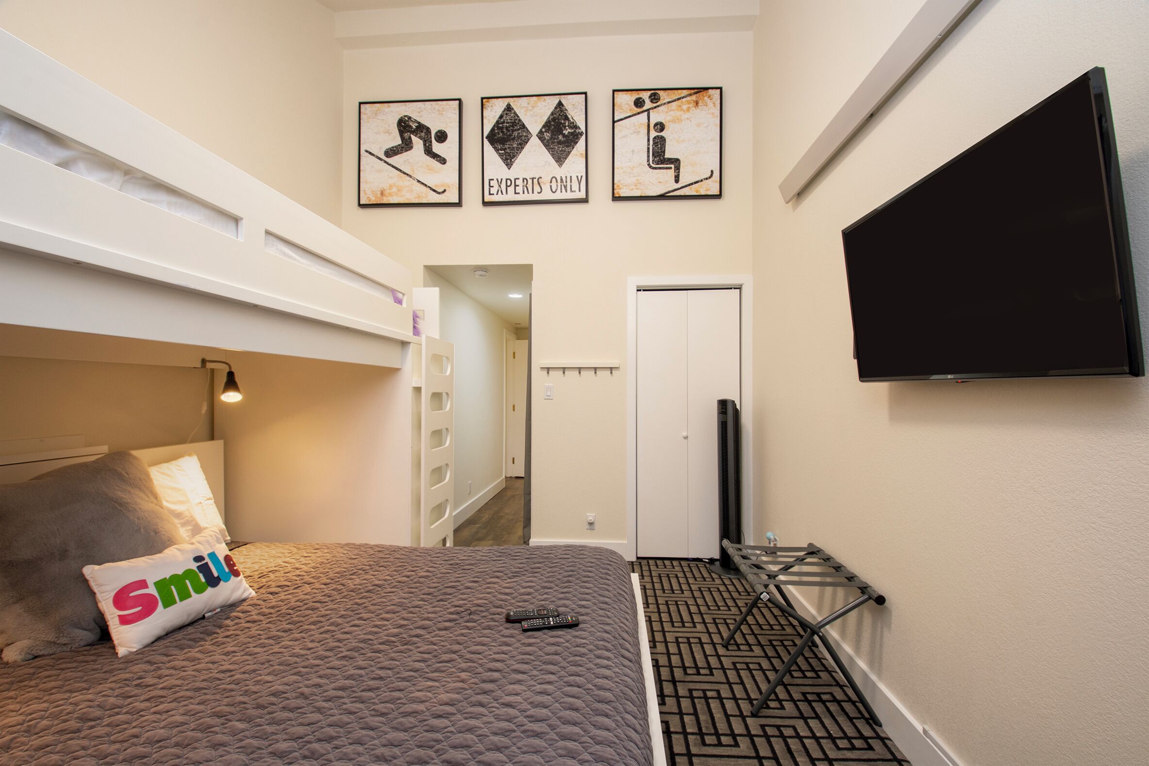 A bedroom with a bunk bed, a bed with a pillow labeled "Smile", a remote control, a TV, and a wall with three framed pictures.