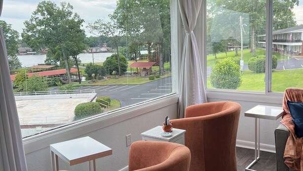 Camera d'albergo con un'ampia finestra che si affaccia su un lago e edifici circostanti. Sono presenti due poltrone arancioni e un piccolo tavolino con un oggetto decorativo.