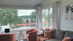Un soggiorno moderno con un divano grigio, poltrone arancioni e un tavolino bianco. Un'ampia finestra offre vista su un lago e degli edifici.