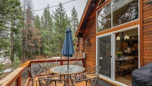 A wooden deck with a glass door leading to a dining area, surrounded by trees.