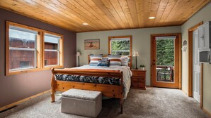 A bedroom with a wooden ceiling, a bed with multiple pillows, a window, a door leading to a balcony, and a small table with chairs.