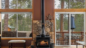 A stone fireplace with a black chimney, a bench, and a table with chairs in a room with large windows and a view of trees.