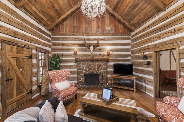 A log cabin interior with a stone fireplace, a television, and a chandelier.