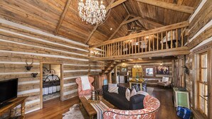 Interior de una cabaña de madera con escalera de madera, candelabro, chimenea y televisor.