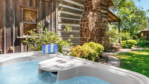 Un jacuzzi frente a una cabaña rústica de troncos con un pilar de piedra, rodeado de follaje.