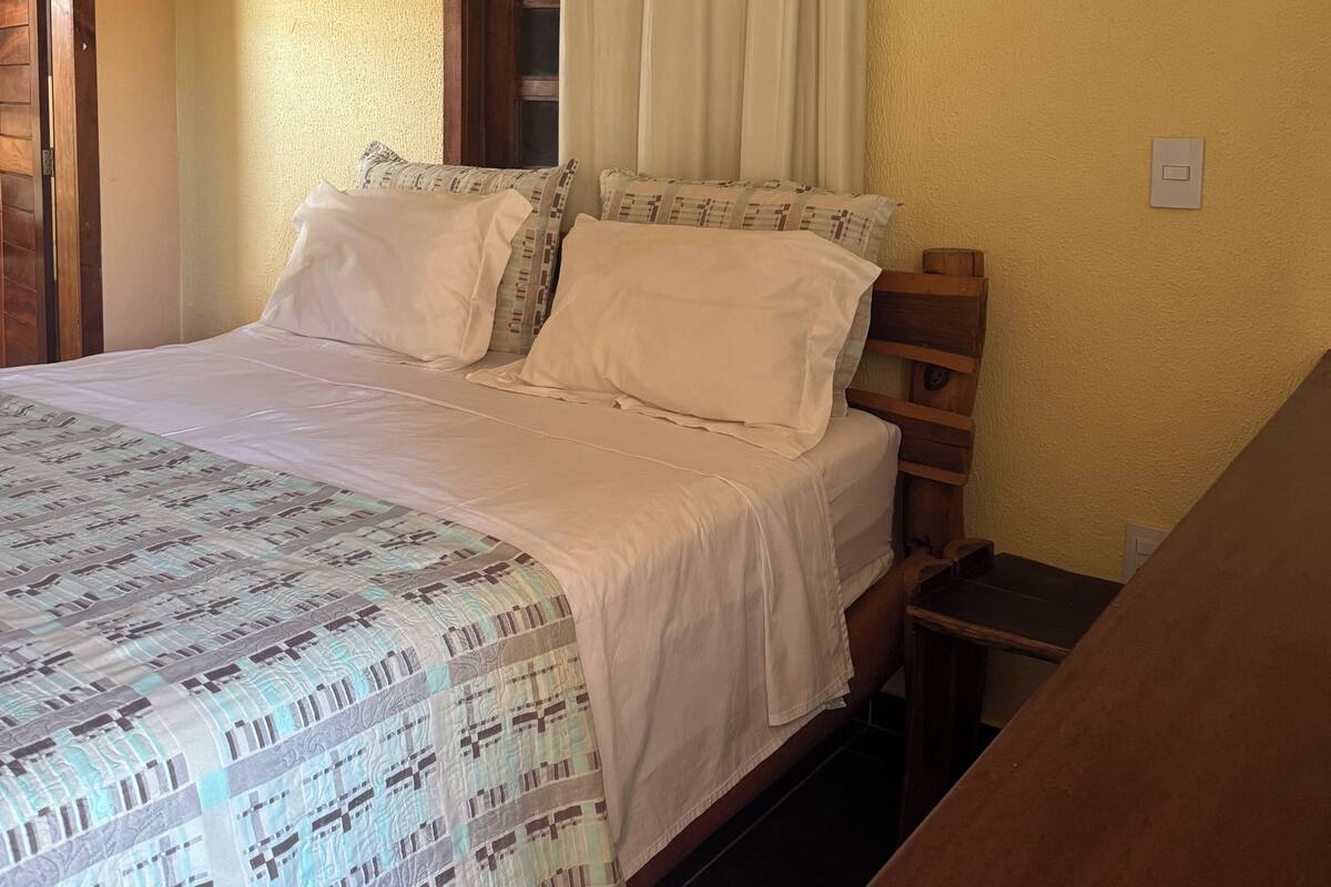 Un dormitorio con cama, cabecera de madera, ventana con cortinas y mesita de noche.