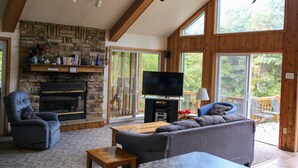 Una sala de estar acogedora con chimenea de piedra, un sofá cómodo, televisión y ventanas amplias que ofrecen vistas al exterior.