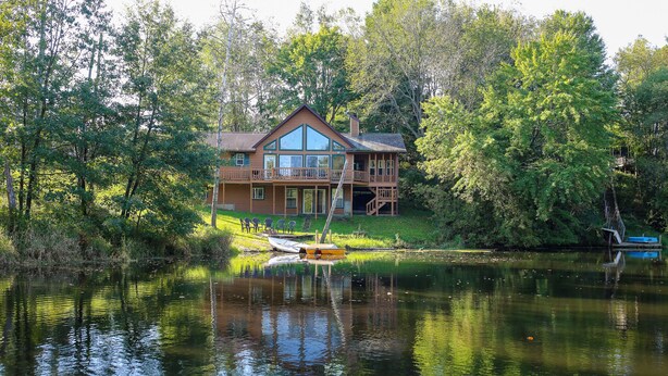 Una casa de madera grande con balcón, ubicada junto a un lago tranquilo rodeado de árboles densos.