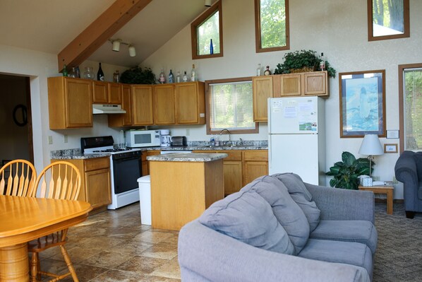 A kitchen with wooden cabinets, a stove, microwave, and a refrigerator. There is a dining table with chairs and a gray sofa in the living area.