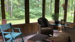 A covered porch with wicker furniture, a blue chair, and a view of a forest. - Quiet and peaceful cottage in Crosslake (Crosslake)