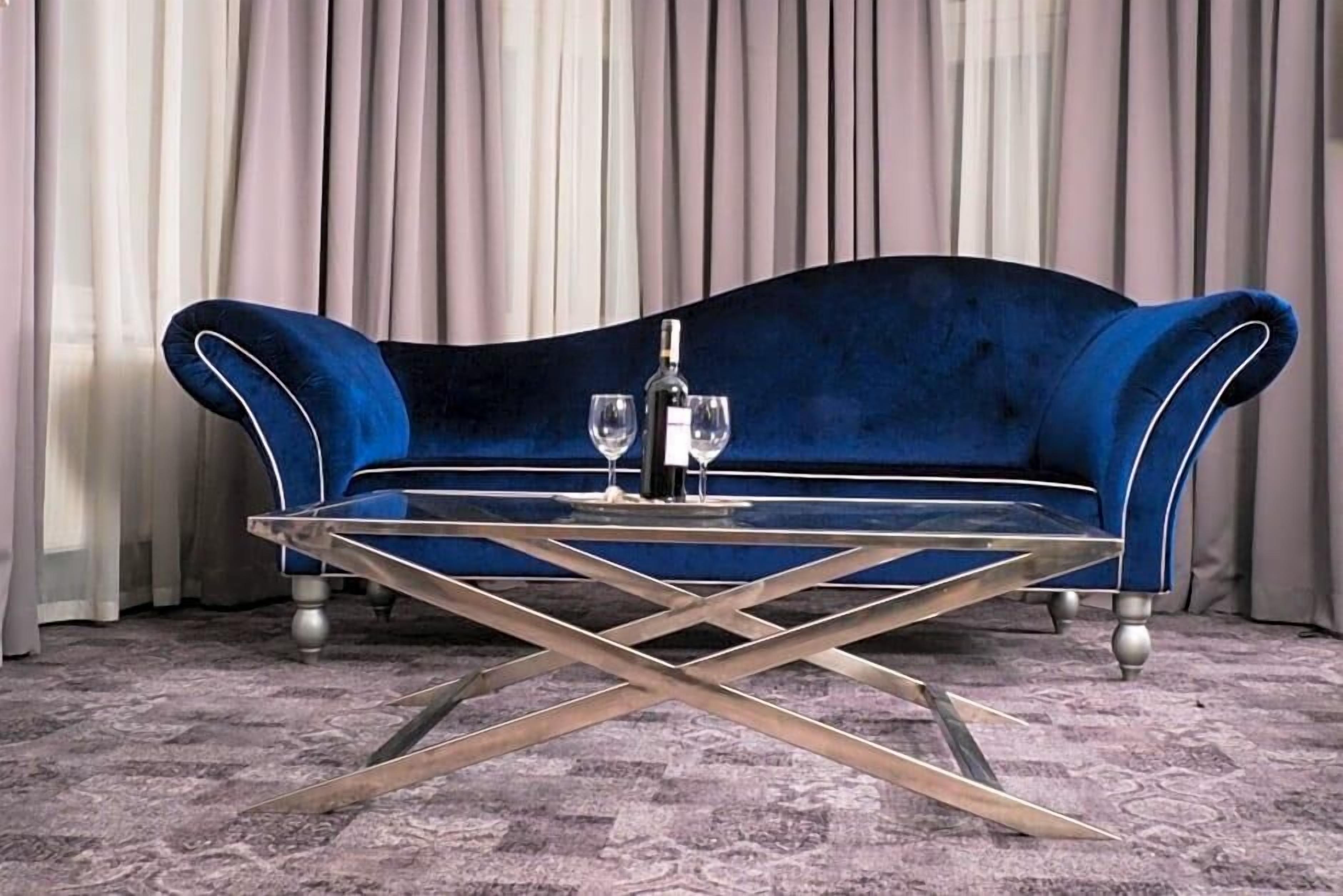A blue velvet sofa with a glass coffee table in front, set against a backdrop of curtains.