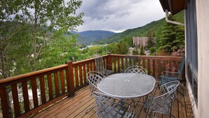 Una terraza de madera con una mesa redonda de metal y cuatro sillas, con vistas a un paisaje montañoso.