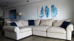 Un sofá seccional color beige con almohadones azules, una lámpara de pie blanca y tres obras de arte con forma de caracola azul en la pared.