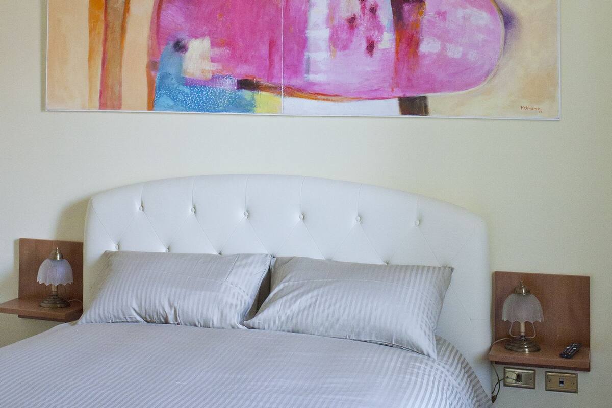Un lit bien fait, avec une tête de lit blanche et deux oreillers, flanqué de deux tables de chevet sur lesquelles reposent des lampes et une petite horloge.