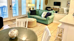A spacious living room with a green sofa, a dining table with chairs, and a kitchen area with a TV. - REDUCED OFF-SEASON RATES!  Oceanfront Condo, Sleeps 4, Close to Everything! (Ocean Isle Beach)