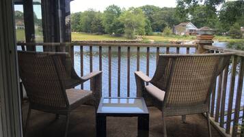 Un balcon avec des fauteuils en osier et une table, d’où l’on peut admirer un plan d’eau et une zone herbeuse, avec des arbres et des maisons en arrière-plan.