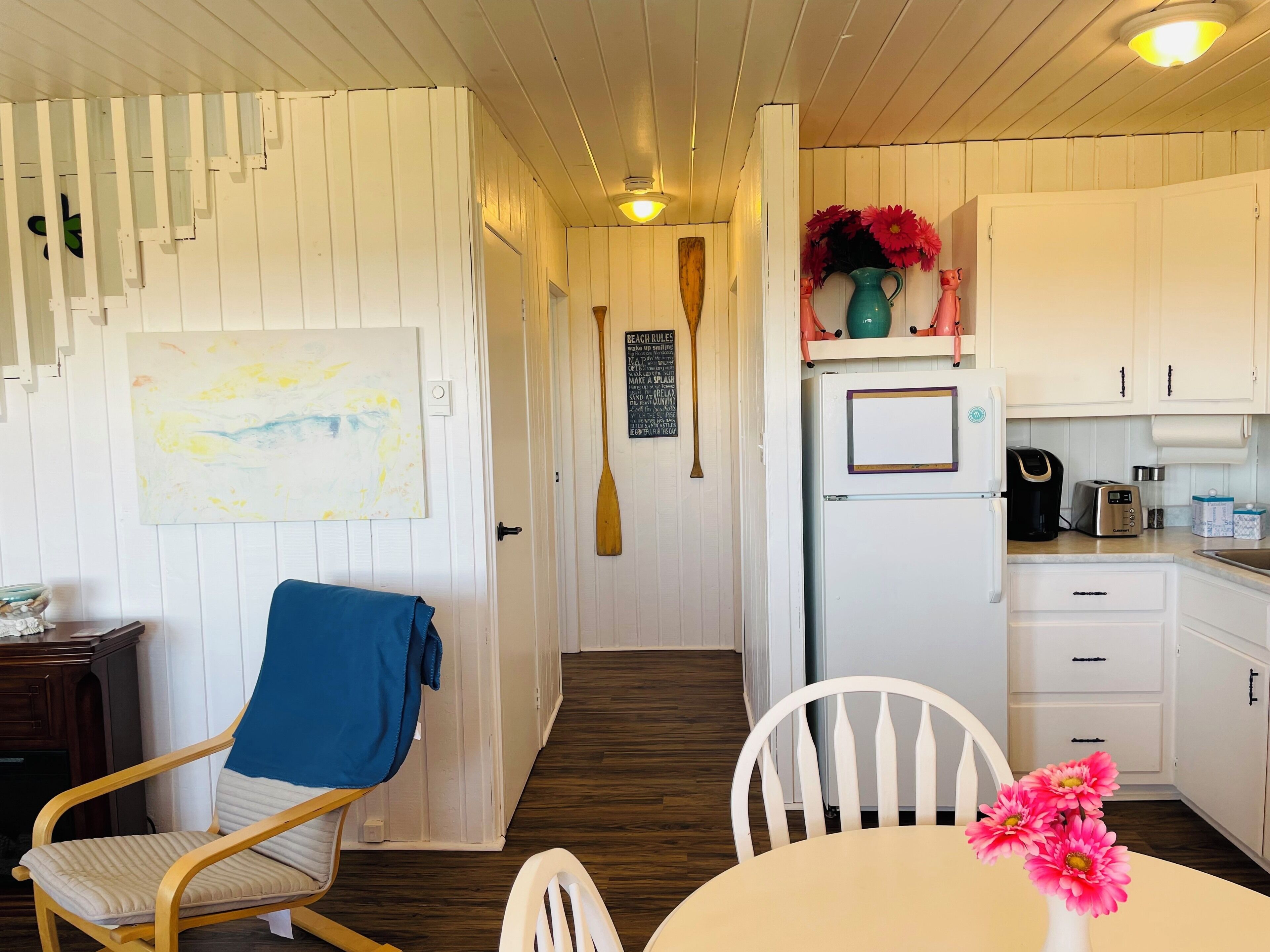 A kitchen with white cabinets, a white refrigerator, a coffee maker, and a toaster. There is a wooden chair with a blue cushion and a white chair with a vase of pink flowers.