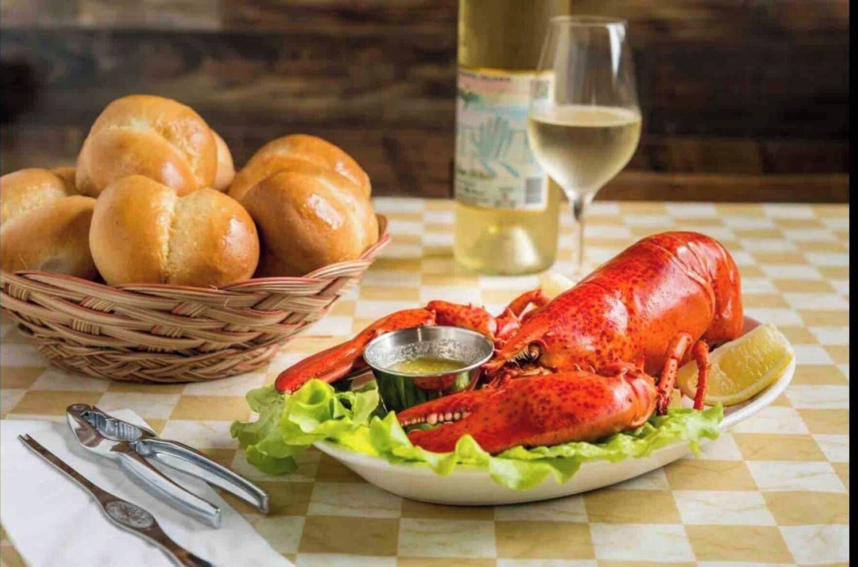 A lobster on a plate with a side of greens and a basket of rolls, a bottle of white wine, and cutlery on a checkered tablecloth.