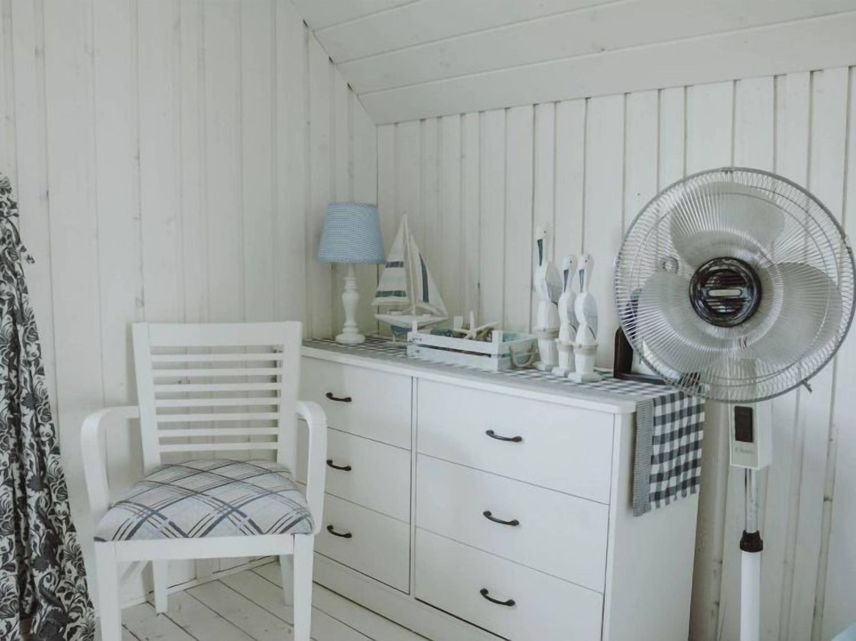Ett vitt rum med en stol, ett litet bord med en lampa, en fläkt och en byrå med lådor.