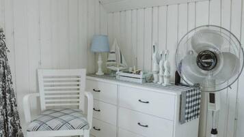 Ett vitt rum med en stol, ett litet bord med en lampa, en fläkt och en byrå med lådor.