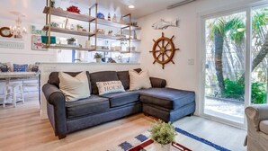 Ein Wohnzimmer mit einem dunkelgrauen Ecksofa, einem Couchtisch mit Pflanze und einem großen Fenster mit Blick auf Palmen.