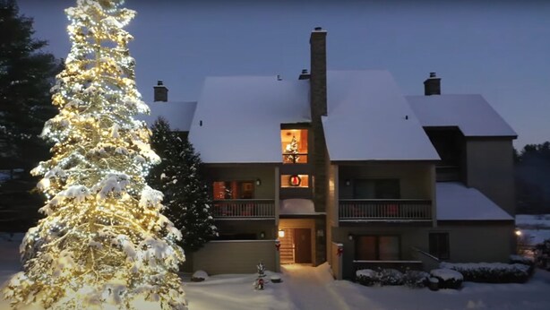 Una casa de dos pisos con tejado cubierto de nieve, un árbol de Navidad iluminado y un balcón con barandillas.