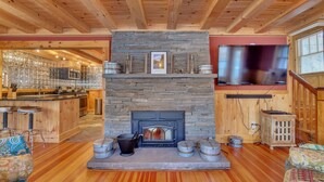 A cozy cabin interior with a stone fireplace, wooden beams, a kitchen area, and a TV mounted on the wall.