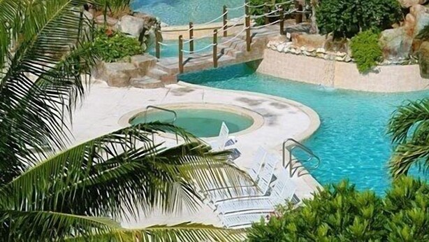 Zona de piscina de un complejo turístico con palmeras, una piscina azul cristalina y vistas al océano.