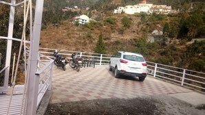 A white SUV parked on a checkered pavement with a white railing, overlooking a hilly area with buildings and trees.