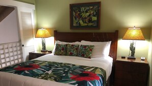Una cama bien tendida con una colcha floral, dos lámparas de noche, una mesita de noche con un reloj y un cuadro en la pared.