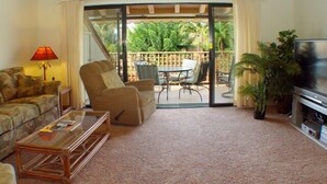 Una sala de estar con sofá, sillón, mesa de centro, televisor y un balcón con mobiliario exterior.